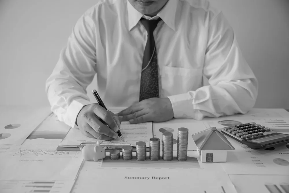 Analyst signing a summary report beside stacked coins and a house model
